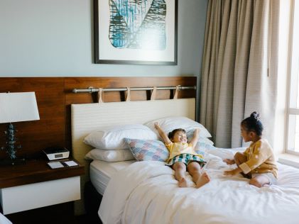 Two children smile while sitting on a comfortable bed in a Suite at The Grand Islander, a Hilton Grand Vacations Club in Oahu, Hawaii