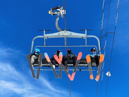 Four skiers look down from a ski lift against a clear blue sky