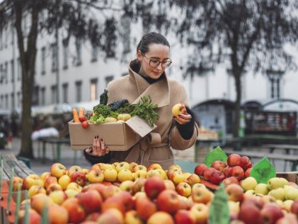 A woman in a coat holding a box of produce at a farmers market