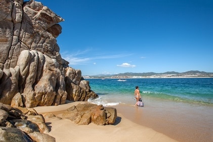 Far shot of woman standing on the shore, facing the water with towering rock formations behind her. She's about to go for a snorkel on a beautifyl blue sky day at Santa Maria Bay, near Los Cabos and San Jose del Cabo, Mexico.