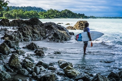 Lone surfer on rocky shore of the Osa Peninsula near Puerto Jiminez in Costa Rica. He is holding a surfboard as he watches the waves beyond.