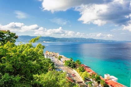 Stunning Montego Bay image of green tropical foliage and the coastline below. Crystal clear waters lie beyond the coastal homes, and In the ar distance are soft mountainous formations, blue skies, and fluffy white cumulous clouds.