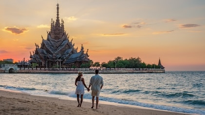 Couple walking along beach in Pattaya Thailand as the Sanctuary of Truth temple is seen in the distance, highlighted by a setting sun.