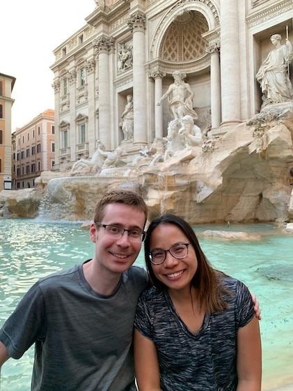 Candid photo of honeymooners in front of the blue waters and white marble figures adorning the Trevi fountain in Rome, Italy.