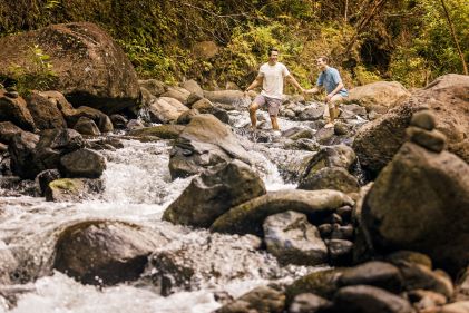 Lush tropical scene, couple crossing rainforest stream, smiling, assisting each other, Iao Valley State Park, Maui, Hawaii. 