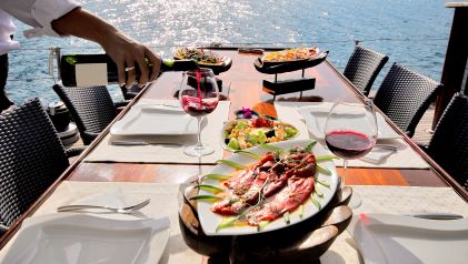 Delicious alfresco spread, set dining table, server pouring wine, sun shining on calm waters.