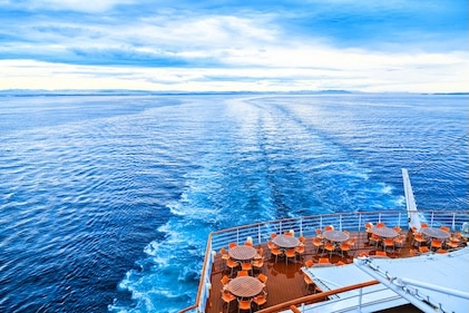 View of cruise ship's large wake on deep blue seas under blue sky with white clouds. Partial view of the stern with dining tables.