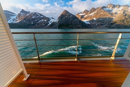 View from cruise ship cabin facing outward onto balcony with ocean then late-day sun shining on snow-capped mountains in the distance.