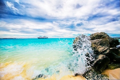 Image with golden sand in the foreground, black rocks with a wave crashing against it, cerulean seas with a cruise ship in the distance under deep blue skies with white clouds.