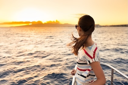 Woman watching the orange glow of a sunset in the distance as she stand on the tip of a ship's bow.